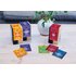 In the picture you can see three Happiness Is tea packs in the colors purple, red and yellow. These are standing on a table. Behind them you can see a plant.